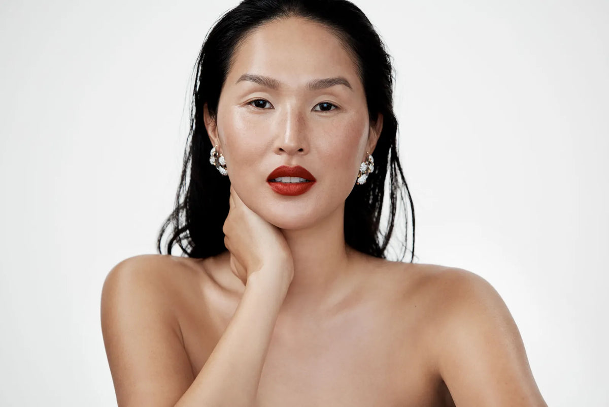 A woman with glowing skin, red lipstick and pearl earrings, her hand at her neck.